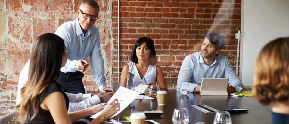 A diverse group of office professionals in a serious meeting around a boardroom table with a brick wall background. A senior male leader is standing and pointing while speaking, as colleagues look on with expressions of concern or focused attention, illustrating a high-pressure workplace environment.