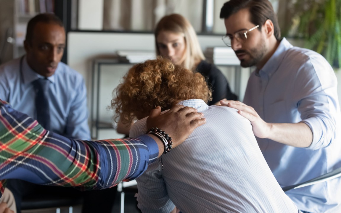 A group of concerned colleagues offering support to a distressed woman who has her head bowed, with two co-workers placing comforting hands on her shoulders in a collaborative office setting.