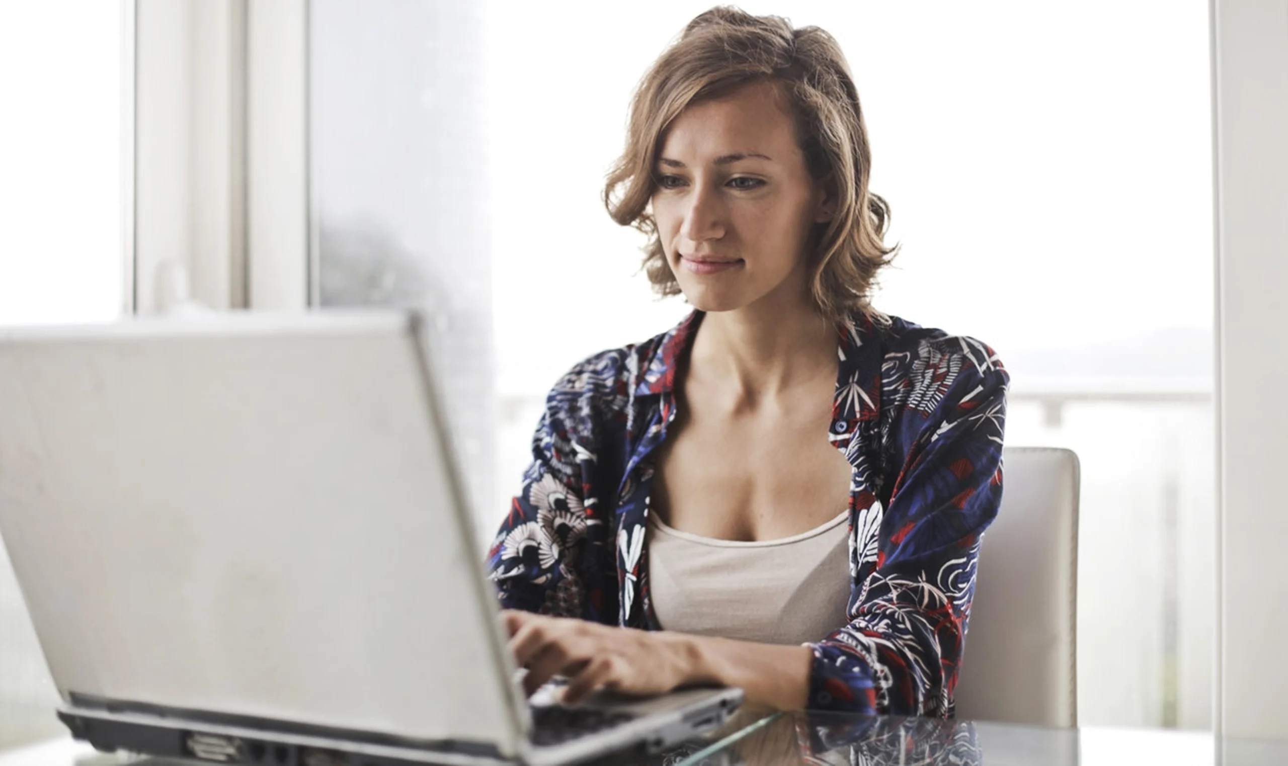 A woman sitting at a glass table, calmly focused on her laptop screen in a bright, naturally lit room.