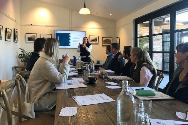 A diverse group of business leaders seated at a conference table for the Future Fit Leaders Program. Emily Johnson speaks in front of a monitor showing the workshop agenda. The setting is a collaborative workshop environment focused on leadership development and mental fitness.