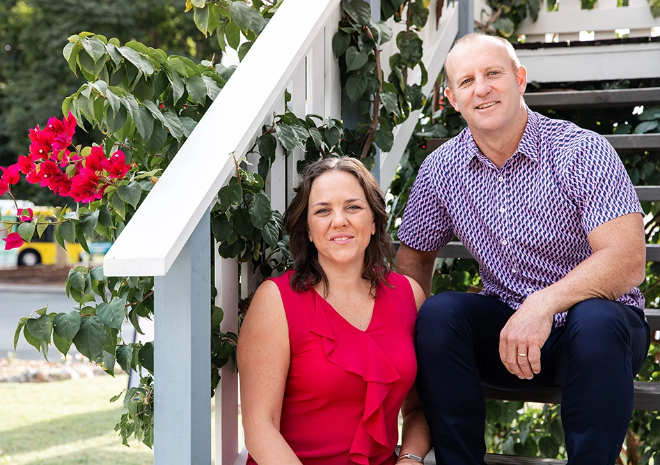 Co-founders Emily and Scott Johnson of Get Mentally Fit sitting together on outdoor stairs, smiling warmly in a professional portrait surrounded by green foliage and red flowers.