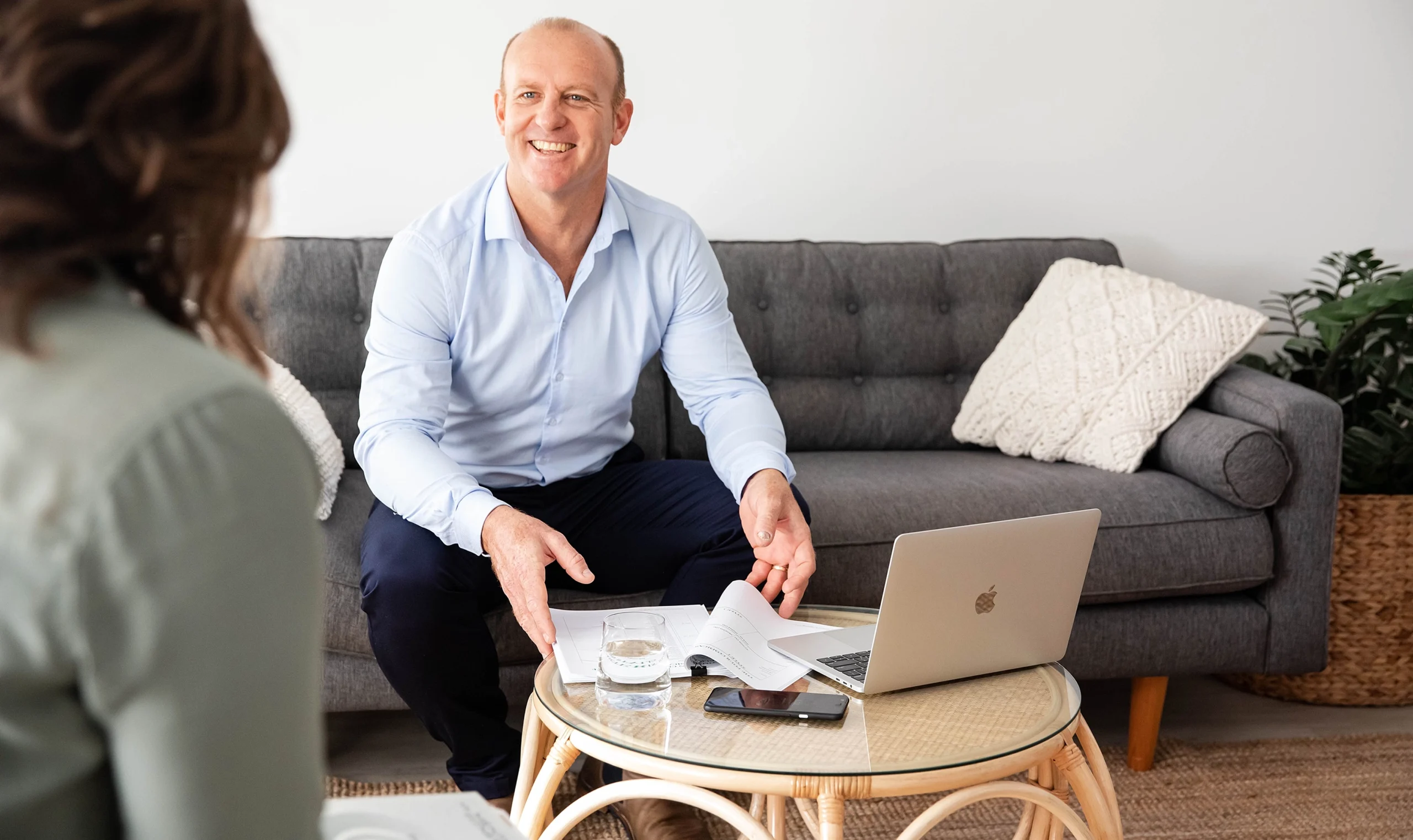 A male professional in a light blue shirt smiling during Get Mentally Fit's Resilience Test consultation, seated on a grey sofa with a laptop and assessment documents on a coffee table.