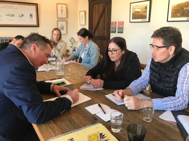 Professionals collaborating during a self-management workshop. A man in a blue suit writes in a workbook while colleagues discuss strategies around a wooden conference table in a bright, modern training room.