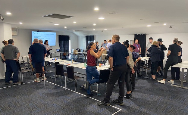 A large group of diverse professionals participating in an interactive social awareness workshop. Participants are standing and discussing tasks in a spacious training room with a presentation screen displaying an organisational definitions exercise.
