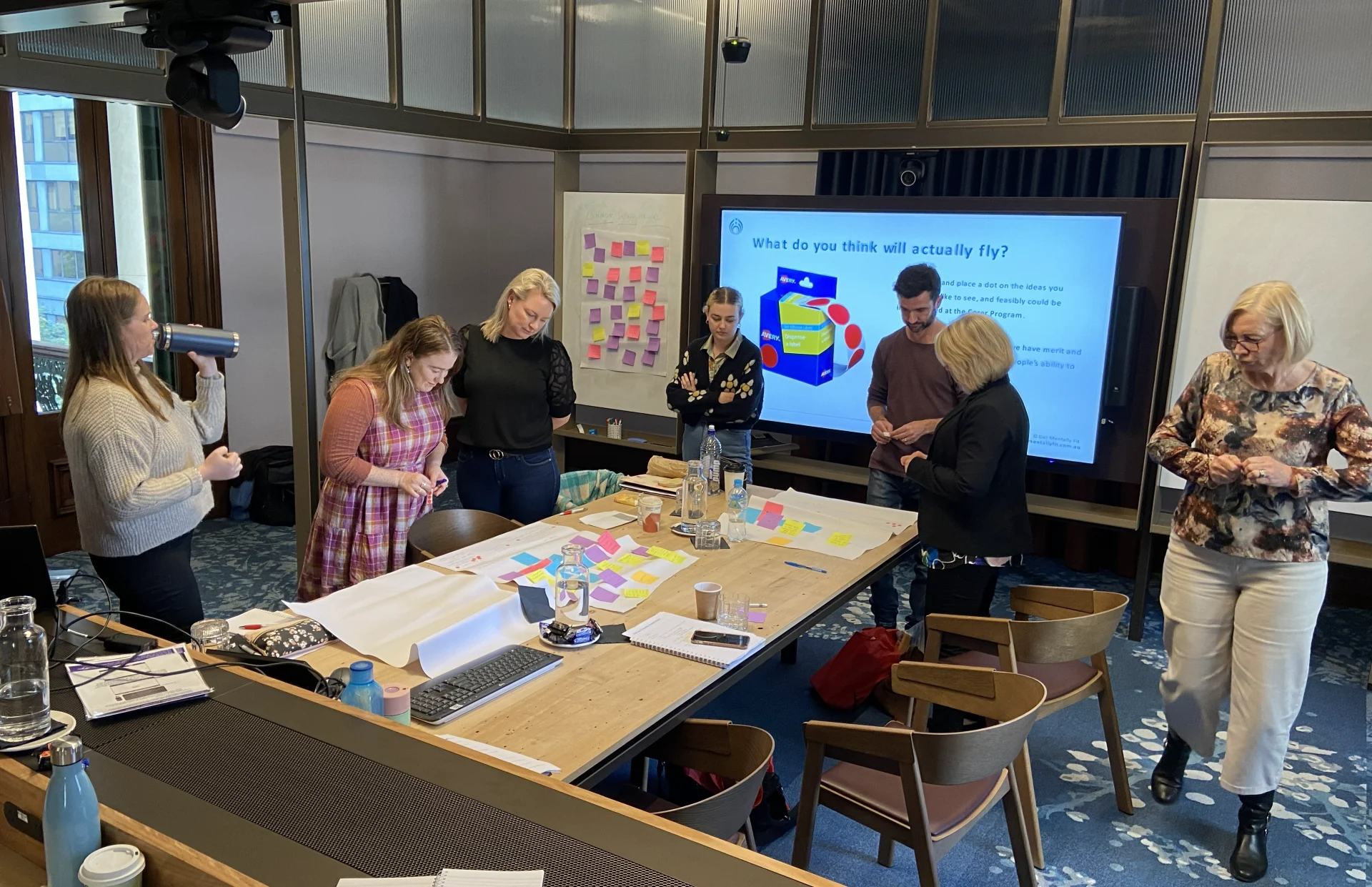 Business professionals participating in a social management and ideation workshop. They are standing around a large table covered in post-it notes and flipchart paper, with a presentation screen in the background asking "What do you think will actually fly?".