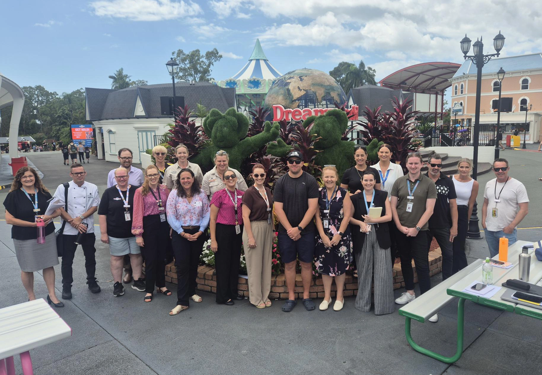 A professional group photo of approximately 20 employees and leaders from multiple departments standing on a stone pathway at Dreamworld, Queensland, with the park’s iconic carousel and globe in the background.