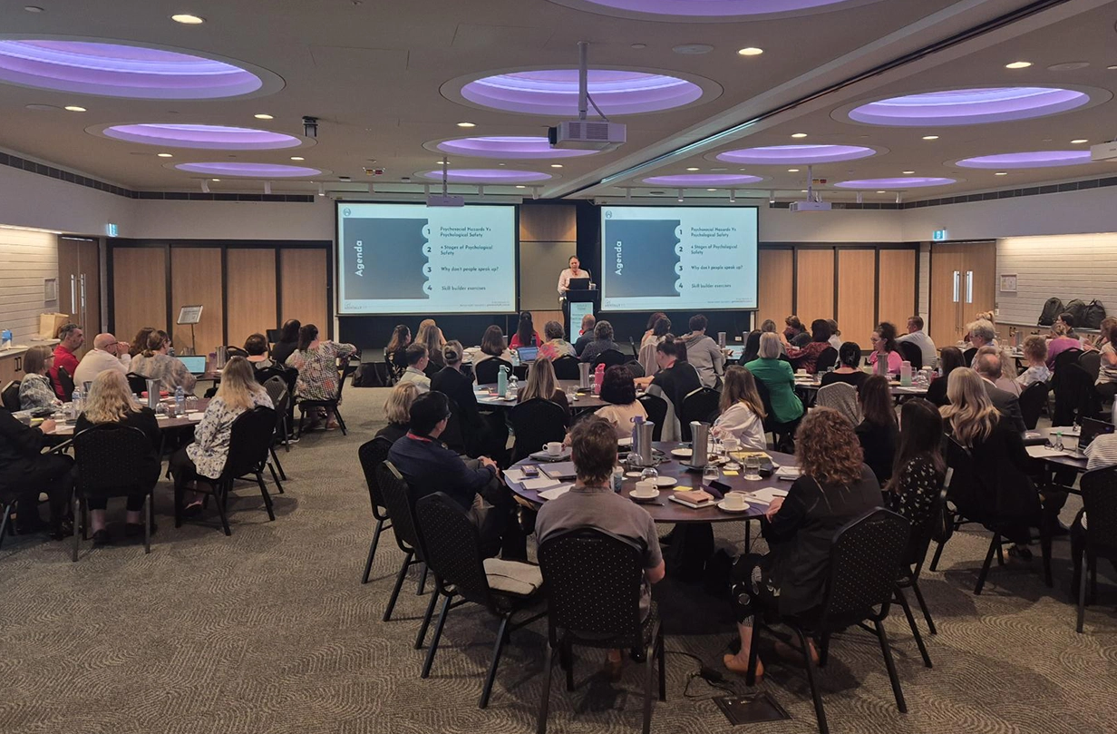 A large group of professionals seated at round tables in a modern conference hall, participating in a staff training workshop with a presenter and dual projector screens displaying an agenda.